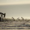 A field with oil pump jacks surrounded by greenery under a cloudy sky and sunlight